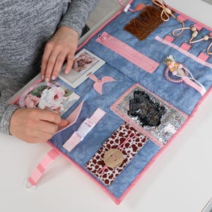 以下が含まれることがあります： さまざまなテクスチャと留め具を備えた感覚アクティビティボード。ボードは青色でピンクのトリムが付いており、ジッパー、バックル、ボタン、その他の触覚要素が特徴です。フォトフレームと装飾的なアクセントが含まれています。