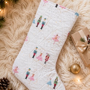 May include: A white quilted Christmas stocking featuring a repeating pattern of nutcracker soldiers and ballerinas in pink, blue, and red outfits. The stocking has a diamond-stitched texture and a white fabric loop at the top for hanging purposes.
