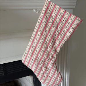 May include: A quilted Christmas stocking featuring a repeating pattern of vertical red and green stripes interspersed with small red heart motifs on a cream-colored background, suspended by a white ribbon loop at the top opening.