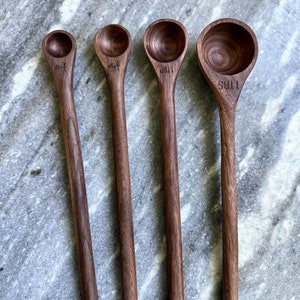 A beautiful set of handcrafted walnut measuring spoons with long, elegant handles, laid out on a marble surface. The rich, dark wood grain contrasts strikingly with the background.
