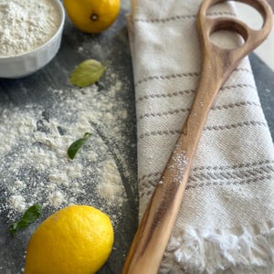 Pu&ograve; includere: Una spatola per impasto in legno con due ritagli circolari e un lungo manico poggia su un canovaccio a righe. Due limoni, una ciotola di farina e foglie di basilico sono sparsi su una superficie scura, suggerendo un tema di cottura.