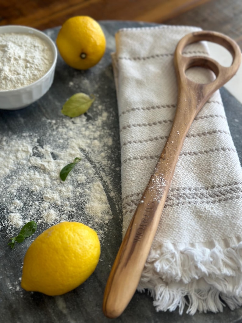 Pu&ograve; includere: Una spatola per impasto in legno con due ritagli circolari e un lungo manico poggia su un canovaccio a righe. Due limoni, una ciotola di farina e foglie di basilico sono sparsi su una superficie scura, suggerendo un tema di cottura.