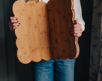 May include: A light brown, cloud-shaped bamboo cutting board. The board is held open, revealing the natural wood grain. The edges have a scalloped design. The board is approximately 18 inches wide and 12 inches tall.
