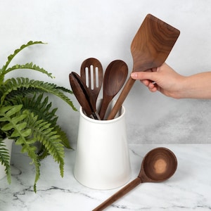 May include: A collection of wooden kitchen utensils, including a spatula, slotted spoon, and tongs, displayed in a white ceramic container. One utensil is being held by a hand. The utensils are made of dark wood and rest on a marble surface.