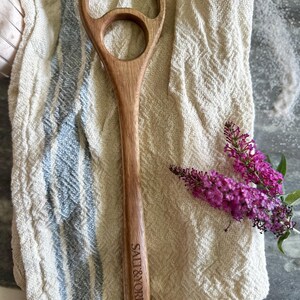 Pu&ograve; includere: Un cucchiaio di legno dal design unico, con una grande apertura circolare e una pi&ugrave; piccola. Il cucchiaio &egrave; inciso con "SALT & YORK" sul manico. &Egrave; appoggiato su un panno color crema a righe blu, accanto a un rametto di fiori viola.