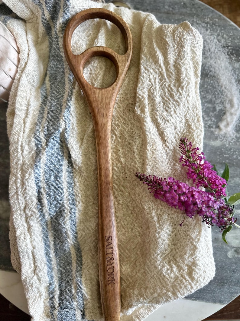 Pu&ograve; includere: Un cucchiaio di legno dal design unico, con una grande apertura circolare e una pi&ugrave; piccola. Il cucchiaio &egrave; inciso con "SALT & YORK" sul manico. &Egrave; appoggiato su un panno color crema a righe blu, accanto a un rametto di fiori viola.