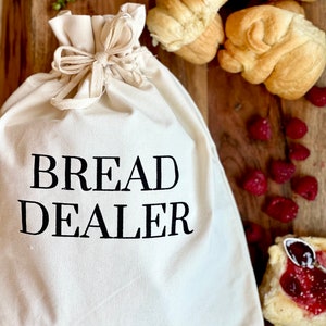 May include: A cream-colored canvas bread bag with the words "BREAD DEALER" printed in black. The bag is tied with a natural-colored drawstring. Freshly baked bread rolls, raspberries, and a pastry are arranged on a wooden surface.