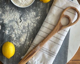 May include: Overhead shot of a wooden spoon, white bowl of flour, two lemons, and a white and gray striped towel on a gray and white marble surface. The spoon has a unique handle design. The scene suggests baking or cooking.
