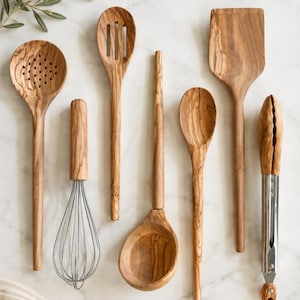May include: A collection of wooden kitchen utensils, including a slotted spoon, whisk, spatula, ladle, and tongs. The utensils are made of light brown wood and are arranged on a white marble surface. The utensils are for cooking and food preparation.
