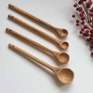 May include: Four wooden measuring spoons with long handles and rounded bowls. Each spoon is engraved with a measurement: 1 TBS, 1 TSP, etc. The spoons are light brown and arranged diagonally. A sprig of red berries is in the background.