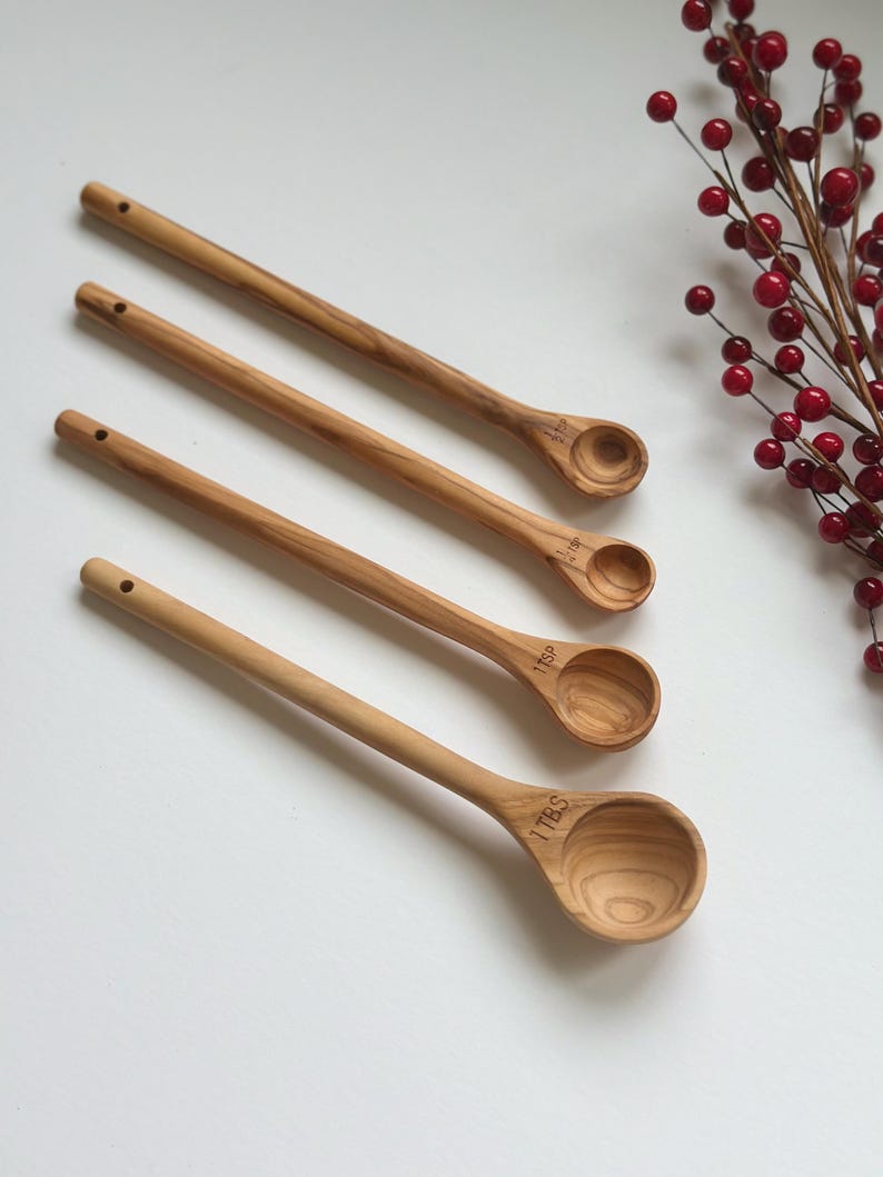 May include: Four wooden measuring spoons with long handles and rounded bowls. Each spoon is engraved with a measurement: 1 TBS, 1 TSP, etc. The spoons are light brown and arranged diagonally. A sprig of red berries is in the background.