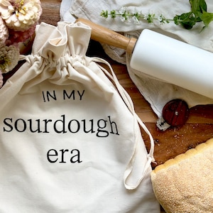 May include: A cream-colored drawstring bag with the text "IN MY sourdough era" in black. A white rolling pin with a wooden handle rests on a linen cloth. A loaf of bread and flowers are also in the image.