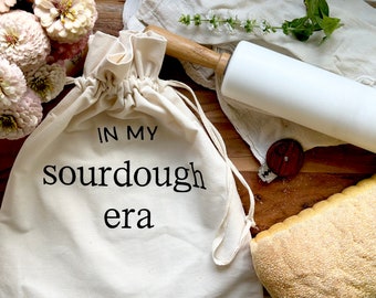 May include: A cream-colored cotton drawstring bag featuring the black printed text "IN MY sourdough era" on the front. The bag is positioned next to a white rolling pin with wooden handles, a small wooden bread lame, and a loaf of bread.