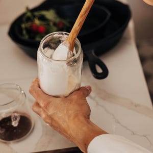 May include: A clear glass jar filled with white powder is held by a person. A wooden spoon is inside the jar, stirring the contents. A black cast iron pan and other jars are in the background. The scene is set on a white countertop.