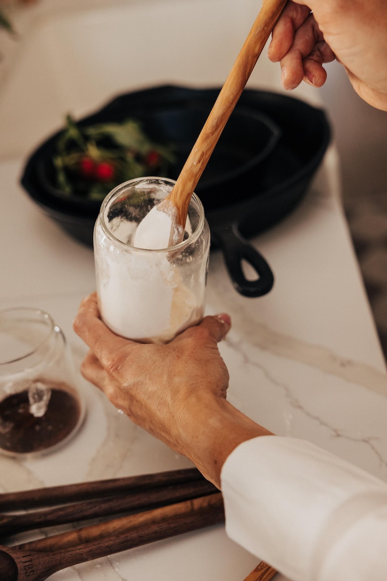May include: A clear glass jar filled with white powder is held by a person. A wooden spoon is inside the jar, stirring the contents. A black cast iron pan and other jars are in the background. The scene is set on a white countertop.