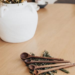 May include: A white ceramic vase with handles holds dried flowers, set on a light wood table. Four wooden measuring spoons rest on rosemary sprigs. The spoons are various sizes, with measurements etched on the handles.