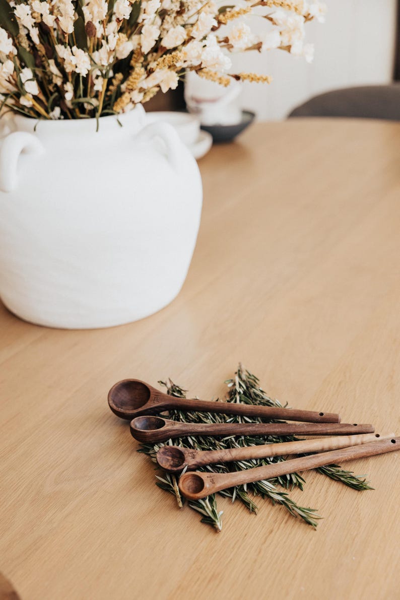 May include: A white ceramic vase with handles holds dried flowers, set on a light wood table. Four wooden measuring spoons rest on rosemary sprigs. The spoons are various sizes, with measurements etched on the handles.