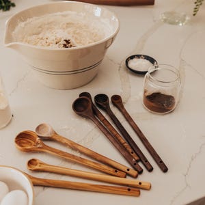 May include: A collection of wooden spoons in various sizes and shades of brown, alongside a large white bowl filled with flour. Other items include a glass jar, a small dish of salt, and a bowl with eggs, suggesting a baking theme.
