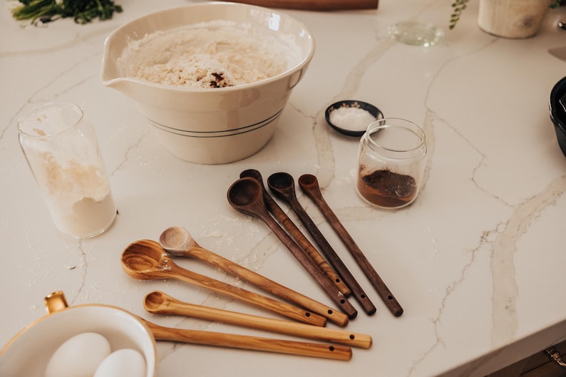 May include: A collection of wooden spoons in various sizes and shades of brown, alongside a large white bowl filled with flour. Other items include a glass jar, a small dish of salt, and a bowl with eggs, suggesting a baking theme.