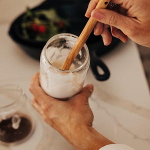May include: A clear glass jar with a white substance inside is being stirred with a wooden spoon. The jar is held by a person's hands. A black pan with green leaves and red berries is in the background.