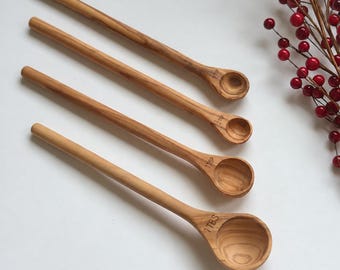 May include: A set of four light brown wooden measuring spoons with long handles. The spoons are labeled 1/4 tsp, 1/2 tsp, 1 tsp, and 1 TBS. The wood features a natural grain pattern, and the spoons are arranged diagonally on a plain surface.