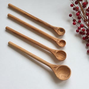 May include: A set of four light brown wooden measuring spoons with long handles. The spoons are labeled 1/4 tsp, 1/2 tsp, 1 tsp, and 1 TBS. The wood features a natural grain pattern, and the spoons are arranged diagonally on a white surface.