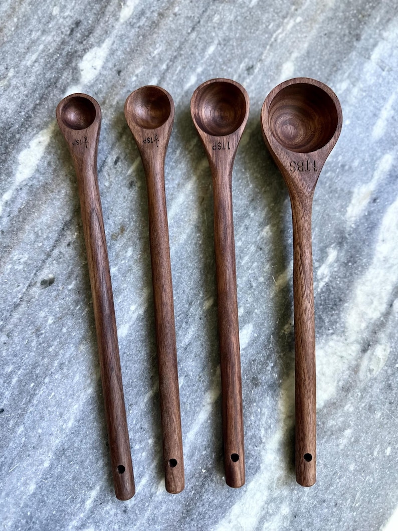 A beautiful set of handcrafted walnut measuring spoons with long, elegant handles, laid out on a marble surface. The rich, dark wood grain contrasts strikingly with the background.