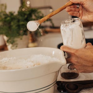 May include: A wooden spoon scoops flour from a glass jar, pouring it into a large white bowl. The bowl has blue stripes and is filled with flour. Other wooden spoons are visible on the counter, suggesting baking or cooking.