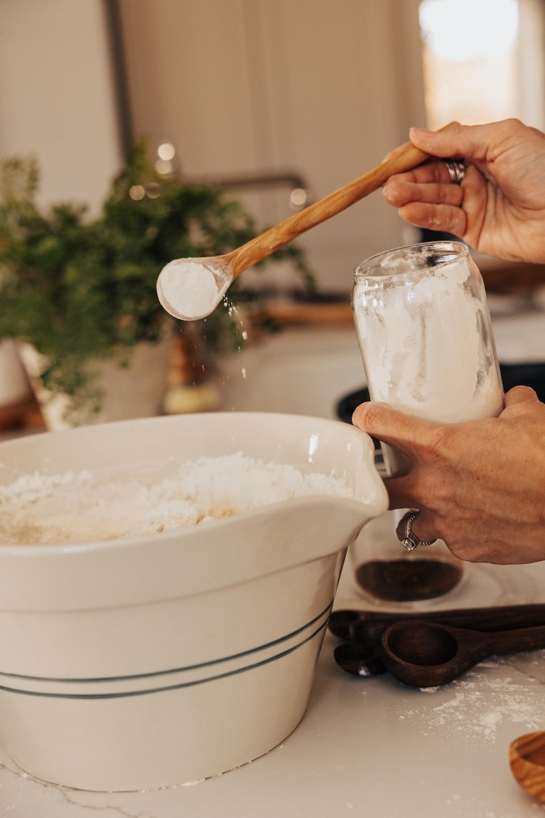 May include: A wooden spoon scoops flour from a glass jar, pouring it into a large white bowl. The bowl has blue stripes and is filled with flour. Other wooden spoons are visible on the counter, suggesting baking or cooking.