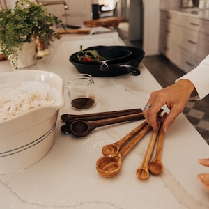 May include: A kitchen scene with various cooking tools. Includes a white bowl with flour, cast iron pans, a small glass jar, and wooden measuring spoons. The spoons are in different sizes and shades of brown.