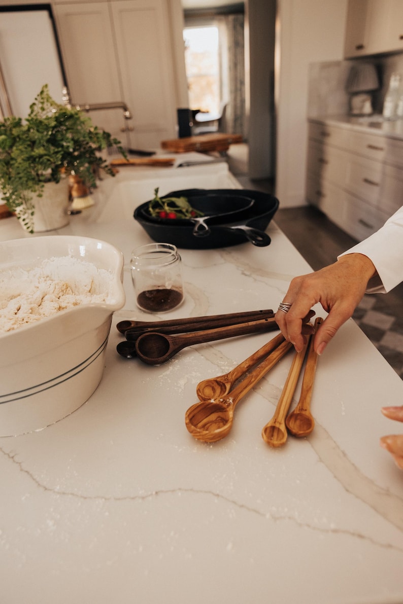 May include: A kitchen scene with various cooking tools. Includes a white bowl with flour, cast iron pans, a small glass jar, and wooden measuring spoons. The spoons are in different sizes and shades of brown.