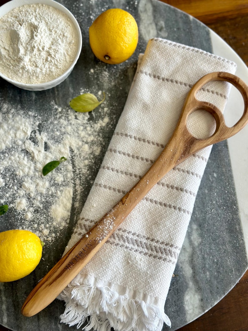 Pu&ograve; includere: Un cucchiaio di legno con un design unico del manico &egrave; appoggiato su un canovaccio a righe bianche e grigie. Sono presenti anche una ciotola di farina, due limoni e un tagliere di marmo. La parola "SALT YORK" &egrave; incisa sul cucchiaio.