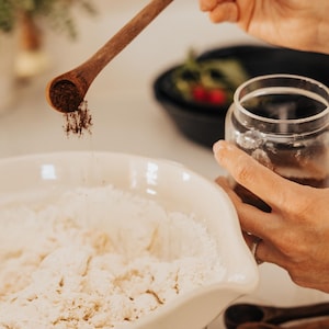 May include: A wooden measuring spoon is held over a white bowl filled with flour, with a clear glass jar in the background. The spoon is filled with a dark powder, likely a spice, being sprinkled into the bowl. The scene suggests baking or cooking.