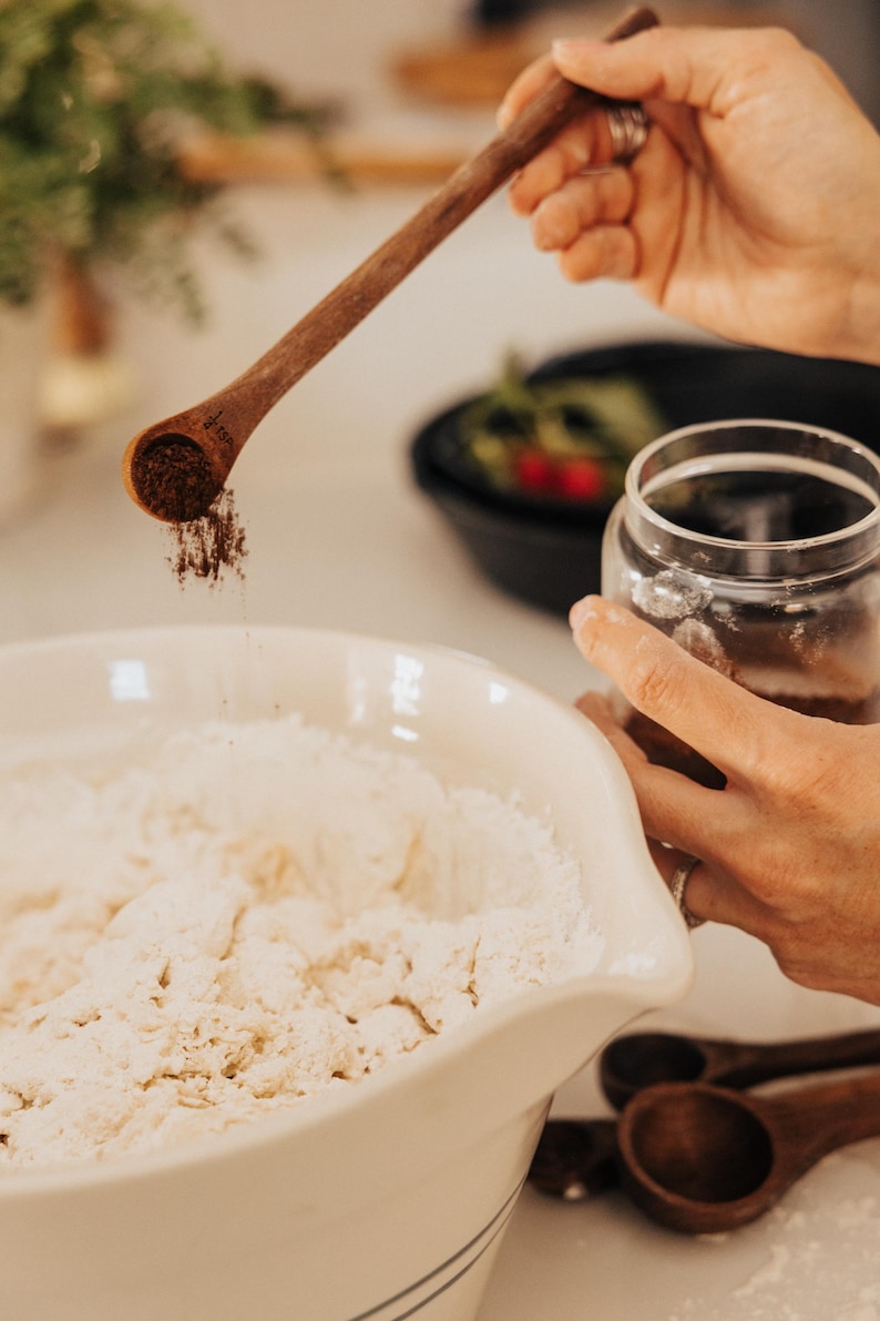 May include: A wooden measuring spoon is held over a white bowl filled with flour, with a clear glass jar in the background. The spoon is filled with a dark powder, likely a spice, being sprinkled into the bowl. The scene suggests baking or cooking.