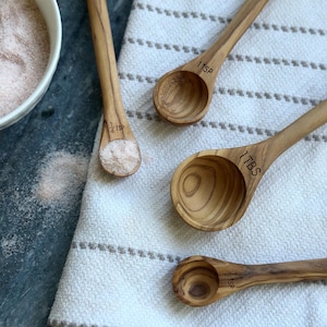 May include: A collection of wooden measuring spoons with engraved measurements. The spoons are made of light-colored wood and are labeled with measurements like "1 TSP" and "1 TBS". One spoon holds a small amount of pink salt. The spoons rest on a white and gray striped towel.