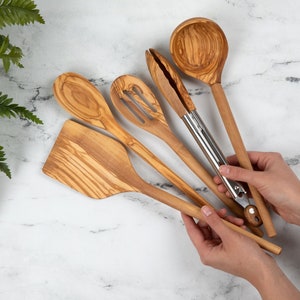 May include: A collection of wooden kitchen utensils, including a spatula, slotted spoon, solid spoon, tongs, and a ladle. The utensils are made of light-colored wood with a natural grain pattern. The tongs have a stainless steel hinge. The background is a white marble surface.