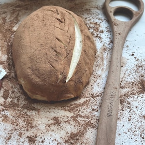 Pu&ograve; includere: Una pagnotta di pane spolverata con polvere marrone, con un'incisione a forma di foglia. Una lama per pane in legno con la scritta "SALT & YORK" &egrave; accanto al pane. Un cestino di vimini &egrave; sullo sfondo.