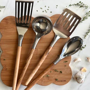 May include: A collection of stainless steel kitchen utensils with wooden handles, including a slotted spatula, ladle, slotted turner, and serving spoon. The utensils are arranged on a wooden cutting board with the text "SALT & YORK".