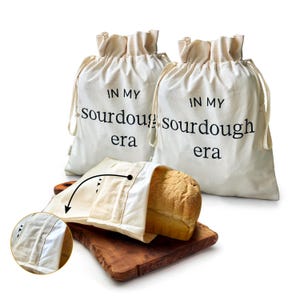 May include: Two off-white drawstring bread bags with the text "IN MY sourdough era" in black. One bag holds a loaf of bread on a wooden cutting board. A close-up shows the interior of the bread bag.