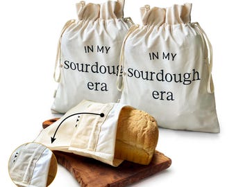 May include: Two off-white drawstring bread bags with the text "IN MY sourdough era" in black. One bag holds a loaf of bread on a wooden cutting board. A close-up shows the interior of the bread bag.