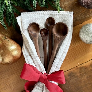 May include: A set of four wooden measuring spoons tied with a red ribbon, presented on a white and beige patterned cloth. The spoons are engraved with measurement labels. Festive holiday decor with gold and silver ornaments and evergreen branches.