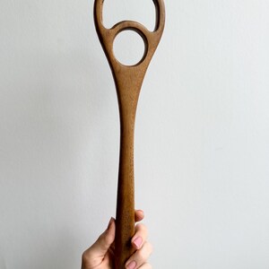 Pu&ograve; includere: Un utensile da cucina in legno con un lungo manico e un design unico. La parte superiore presenta una grande apertura arrotondata con un foro circolare pi&ugrave; piccolo sottostante. L'utensile &egrave; realizzato in legno marrone chiaro e sembra essere un cucchiaio per mescolare.