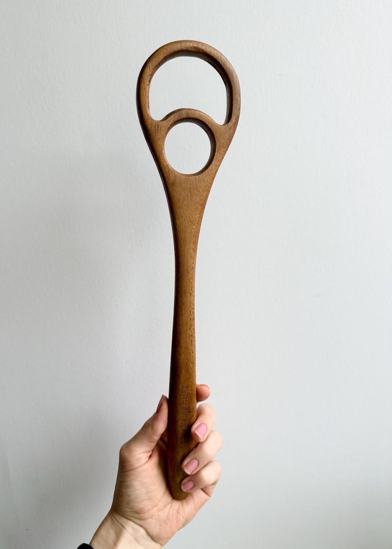 Pu&ograve; includere: Un utensile da cucina in legno con un lungo manico e un design unico. La parte superiore presenta una grande apertura arrotondata con un foro circolare pi&ugrave; piccolo sottostante. L'utensile &egrave; realizzato in legno marrone chiaro e sembra essere un cucchiaio per mescolare.