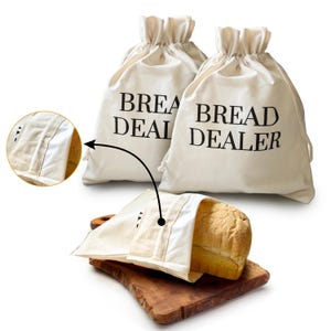 May include: Two off-white drawstring bread bags with the words "BREAD DEALER" printed in black. A loaf of bread is partially visible inside a matching bag, resting on a wooden cutting board. The bags are designed for storing bread.