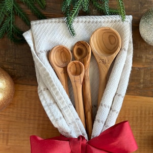 May include: A set of four wooden measuring spoons nestled in a cream-colored cloth, tied with a red ribbon. The spoons are marked with "1 TBS", "1 TSP", and "1/2 TSP". Green pine branches and gold ornaments decorate the background.