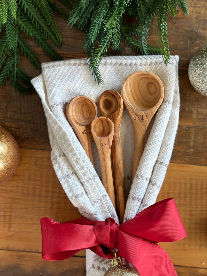 May include: A set of four wooden measuring spoons nestled in a cream-colored cloth, tied with a red ribbon. The spoons are marked with "1 TBS", "1 TSP", and "1/2 TSP". Green pine branches and gold ornaments decorate the background.