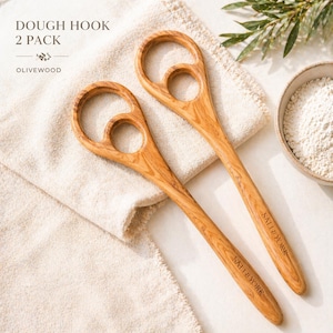 Pu&ograve; includere: Due ganci per impasto in legno d'ulivo con un caratteristico design a manico circolare. Il testo "DOUGH HOOK 2 PACK OLIVEWOOD" &egrave; visibile in alto. I manici sono incisi con "SALT & YORK". I ganci poggiano su un panno beige, con una ciotola di farina sullo sfondo.