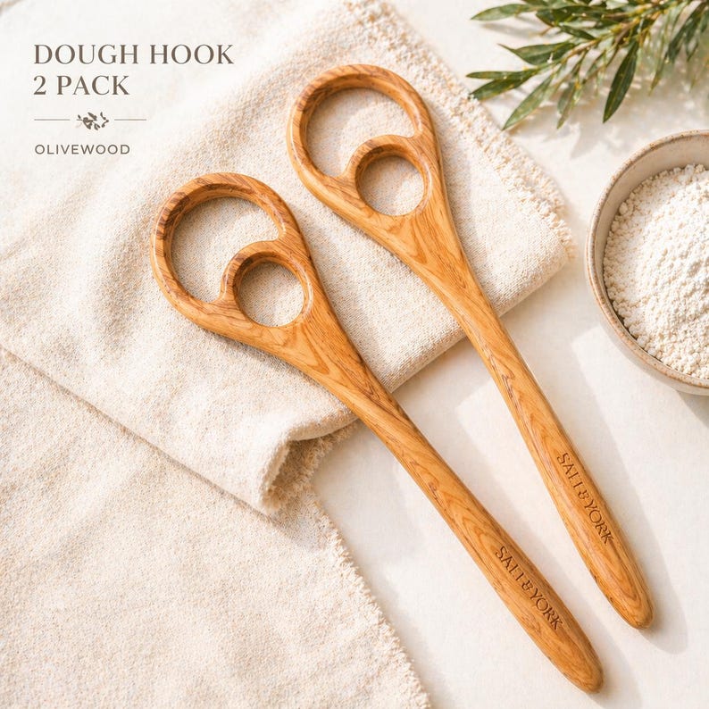 Pu&ograve; includere: Due ganci per impasto in legno d'ulivo con un caratteristico design a manico circolare. Il testo "DOUGH HOOK 2 PACK OLIVEWOOD" &egrave; visibile in alto. I manici sono incisi con "SALT & YORK". I ganci poggiano su un panno beige, con una ciotola di farina sullo sfondo.