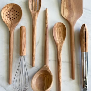May include: A collection of wooden kitchen utensils, including a slotted spoon, spatula, ladle, and whisk. The utensils are made of light-colored wood with visible grain patterns. A metal and wood tong is also included.