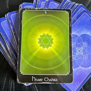 May include: A tarot card deck, with the "Heart Chakra" card in focus. The card has a green and yellow design, a central starburst, and the number "4". The card is labelled "Heart Chakra". The other cards are blue.